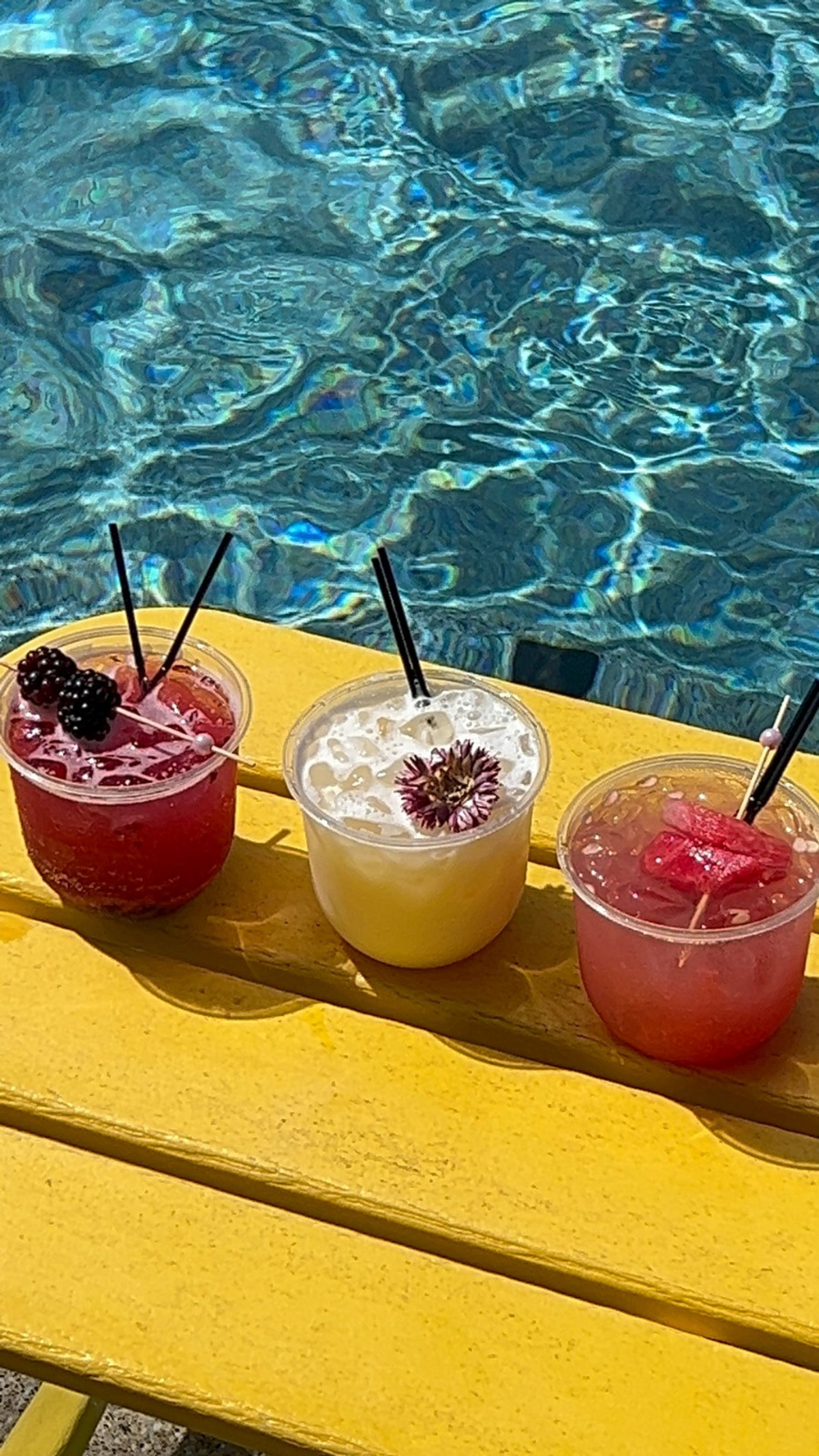 Vibrant poolside trio of cocktails in clear plastic cups with blackberry, edible flower, and watermelon garnishes set on a bright yellow bench beside shimmering blue pool water.