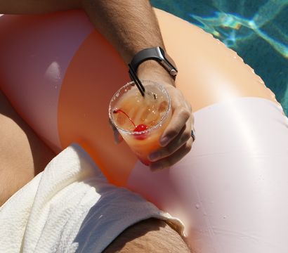 Sunlit poolside scene: arm with smartwatch resting on a peach inflatable float, holding a cherry-topped cocktail over shimmering blue water