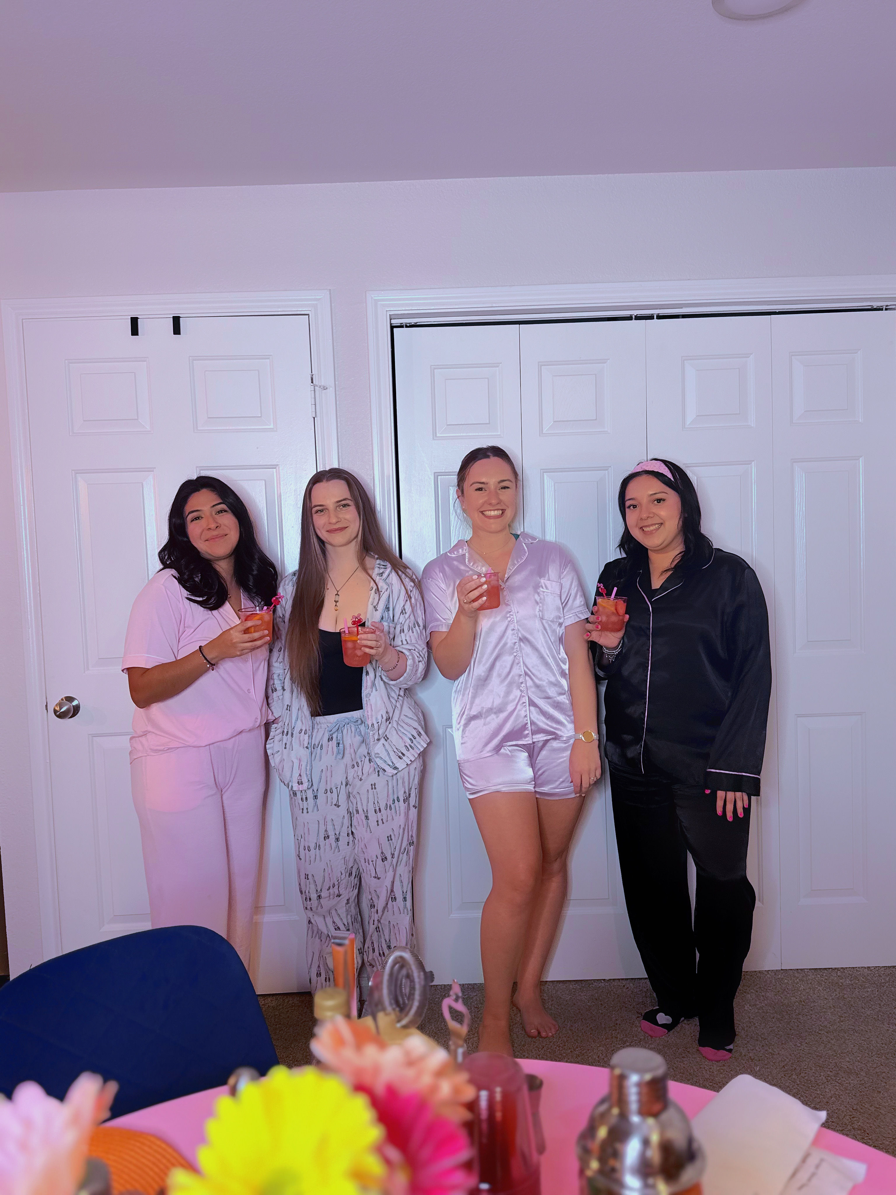 Four friends in pastel and black pajamas holding pink cocktails, smiling and posing in front of white closet doors at a cozy indoor pajama party, with a pink table, bright flowers and drink accessories in the foreground.