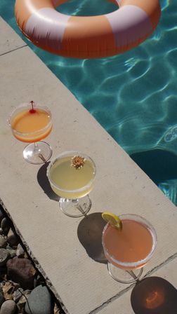 Sunlit poolside scene with three colorful cocktails on a concrete deck beside turquoise water and an orange-and-white inflatable ring, drinks garnished with a cherry, a small flower, and a lemon slice.