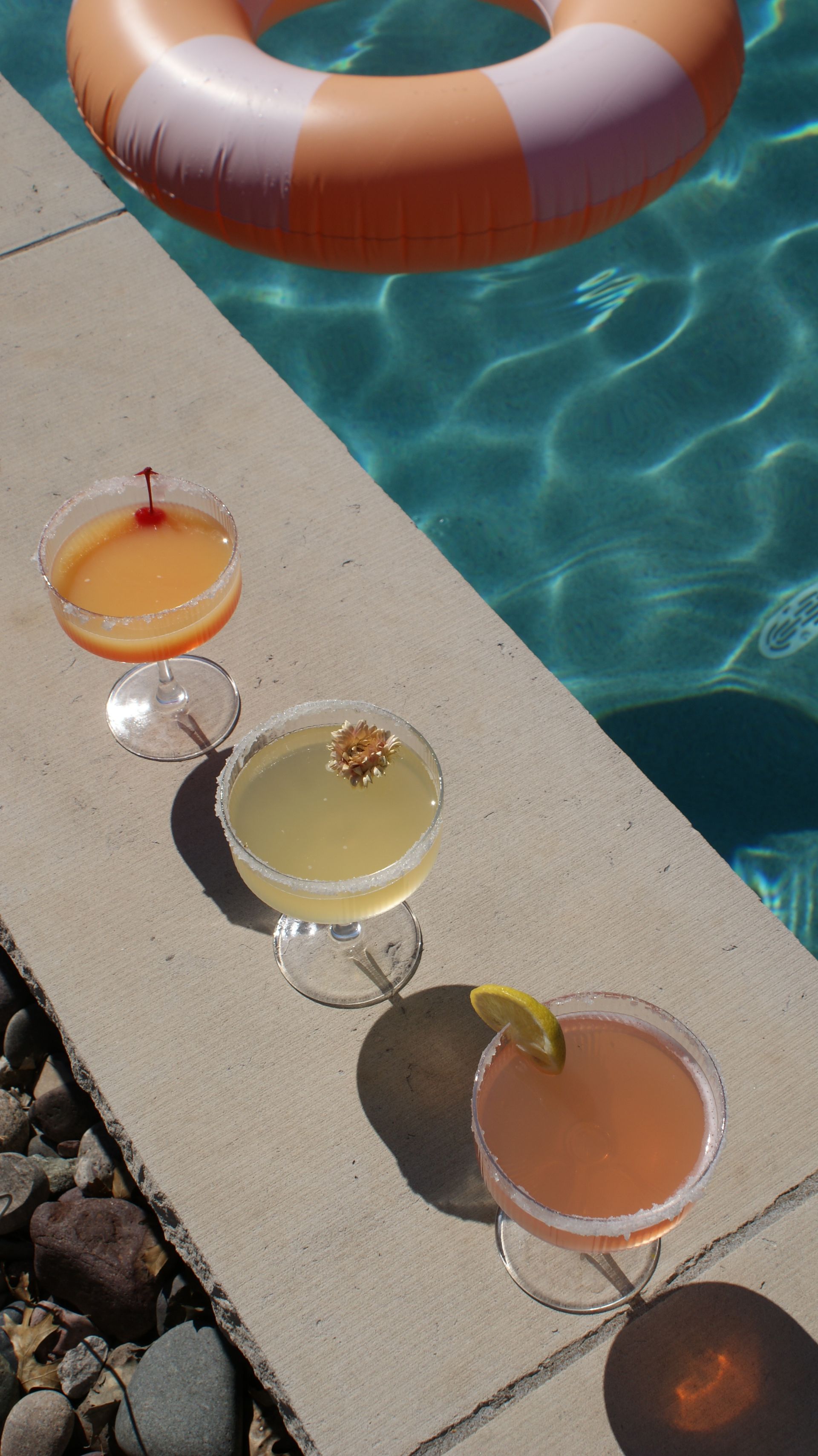 Sunlit poolside scene with three colorful cocktails on a concrete deck beside turquoise water and an orange-and-white inflatable ring, drinks garnished with a cherry, a small flower, and a lemon slice.