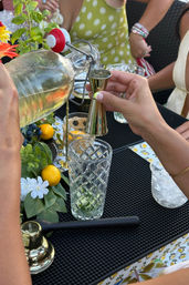 Hands pouring pale yellow liqueur from a swing-top glass bottle into a brass jigger above a tall cut-crystal tumbler with lime, surrounded by small citrus, greenery and flowers on a patterned outdoor patio table at a cocktail gathering.