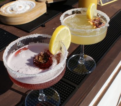 Sunlit outdoor bar counter with two cocktails — a pink floral-garnished drink and a lemon margarita with salted rims and lemon slices, juice bottles in the background for a summer patio vibe.