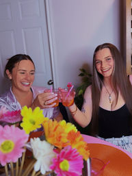 Two friends smiling and toasting with pink cocktails at a table decorated with bright gerbera-style flowers in a cozy indoor setting