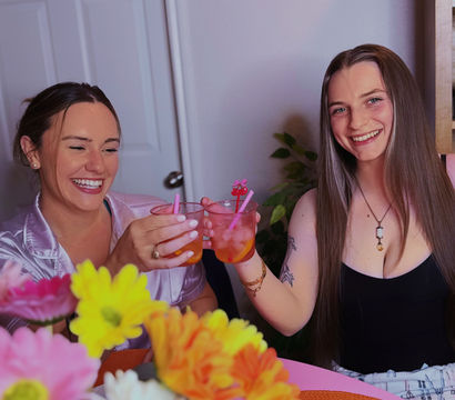Two friends smiling and toasting with pink cocktails at a table decorated with bright gerbera-style flowers in a cozy indoor setting