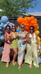 Four friends at a sunny backyard summer party holding cocktails, dressed in colorful summer outfits and posing in front of vibrant blue and orange balloon arches and garden decorations.