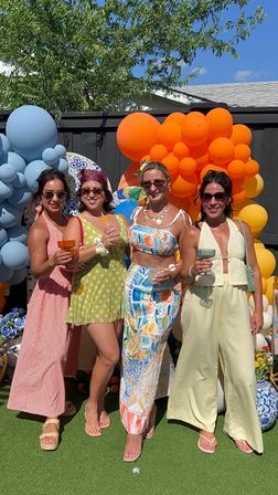 Four friends at a sunny backyard summer party holding cocktails, dressed in colorful summer outfits and posing in front of vibrant blue and orange balloon arches and garden decorations.