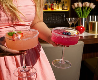 Two vibrant pink cocktails in coupe glasses with sugared rims and fruit garnishes, carried by a server in a pink dress at a bar with bottles and tulips in the background.