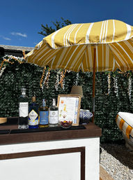Sunny backyard patio bar with a yellow-striped umbrella shading liquor bottles, a framed cocktail menu and a bowl of garnishes on a wooden counter against a leafy hedge under a bright blue sky.