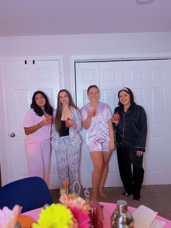 Four friends in colorful pajamas holding pink cocktails at an indoor pajama party in a bedroom, with a pink table, flowers and drinks in the foreground.