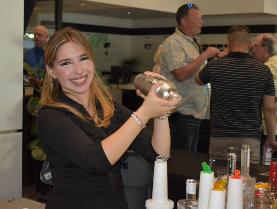 Smiling bartender shaking a metal cocktail shaker at an indoor bar event, with bottles, garnish dispensers and guests chatting in the background.