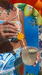 Close-up of a person in a colorful citrus-print summer outfit holding a chilled lime cocktail with an orange-slice straw topper, yellow-and-white nail art, stacked rings and shell necklace against a bright blue lemon-decor backdrop