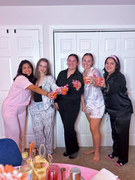 Five smiling friends at an at-home pajama party toasting pink cocktails in front of white closet doors, pink drink setup in foreground.