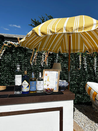 Sunny backyard patio bar under a yellow-striped umbrella with bottles of spirits, framed cocktail menu, floral green wall and bright blue sky