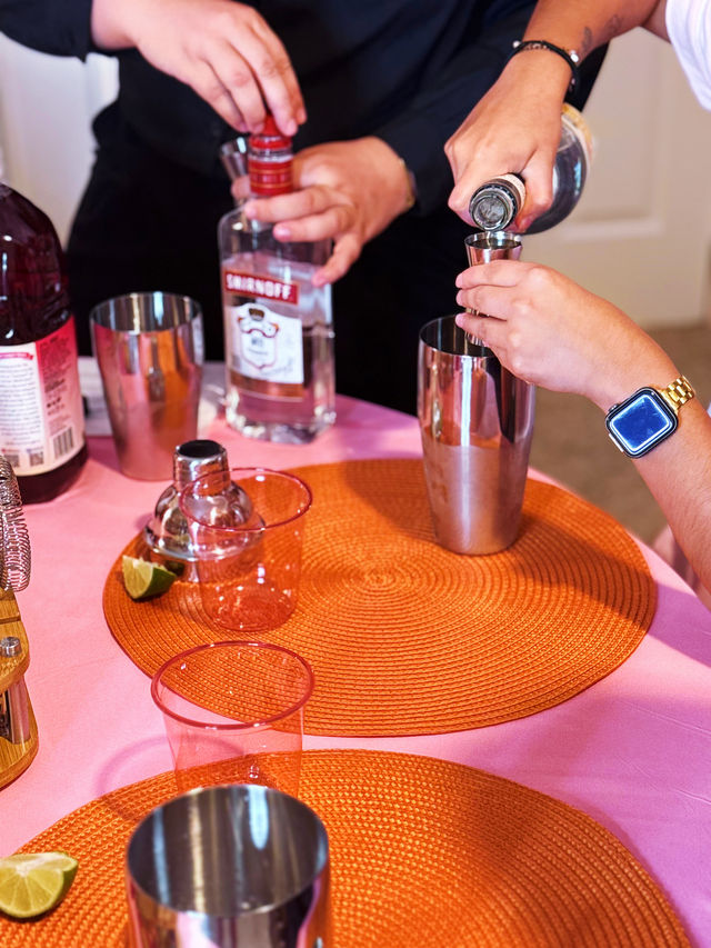 Close-up of hands pouring vodka into a jigger and metal shaker at a vibrant home cocktail station with a pink tablecloth, orange woven placemats, lime wedges and glassware.