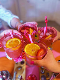 Four hands toasting pink fruity cocktails in translucent cups with orange slices, strawberries, ice and pink straws over a colorful party table — festive summer drinks at a cheerful gathering