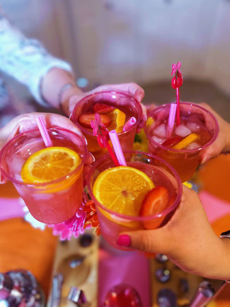 Four hands toasting pink fruity cocktails in translucent cups with orange slices, strawberries, ice and pink straws over a colorful party table — festive summer drinks at a cheerful gathering