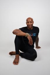 Smiling male yoga practitioner in black athletic wear seated in a seated spinal twist on a white studio background, wearing beaded bracelets — fitness and wellness portrait.
