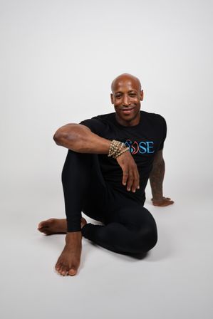 Smiling male yoga practitioner in black athletic wear seated in a seated spinal twist on a white studio background, wearing beaded bracelets — fitness and wellness portrait.