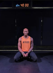 Fit man kneeling on a dark gym floor in an orange tank top and black leggings, smiling beneath a digital 40:00 timer in a studio workout setting.