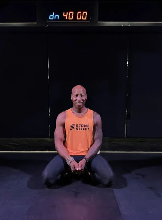 Fit man kneeling on a dark gym floor in an orange tank top and black leggings, smiling beneath a digital 40:00 timer in a studio workout setting.