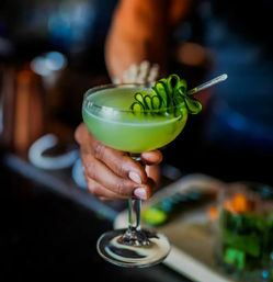 Vibrant lime-green cocktail in a coupe glass with a curled cucumber garnish, held by a hand at a dim cocktail bar.