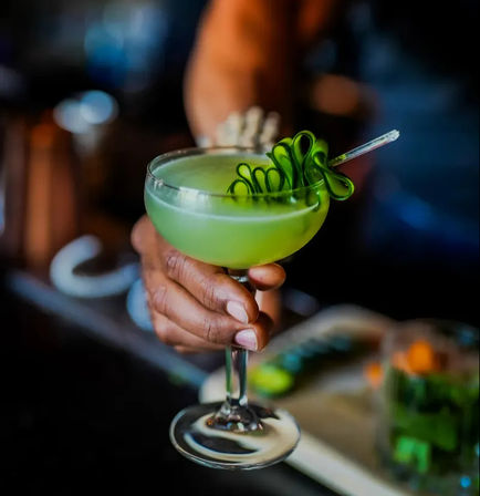 Vibrant lime-green cocktail in a coupe glass with a curled cucumber garnish, held by a hand at a dim cocktail bar.