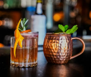 Two craft cocktails on a bar counter: a whiskey-style drink in a rocks glass with an orange twist and rosemary, and a mint-topped Moscow Mule in a hammered copper mug.