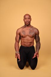 Shirtless muscular fitness athlete kneeling on warm yellow studio background, showing chest and sleeve tattoos, wearing black compression leggings and beaded bracelets, hands extended downward in a calm meditative gesture.