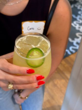 Hand with bright red nails and gold rings holding a pale yellow-green jalapeño cocktail on ice in a stemless glass, person in a black top with a name sticker at an indoor event.