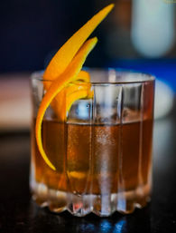 Whiskey cocktail on the rocks in a faceted lowball glass, garnished with a long orange peel twist — close-up on a dimly lit bar surface.