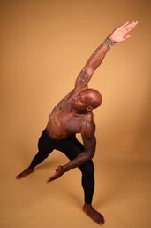 Muscular tattooed man in black leggings performing a side-angle yoga stretch in a warm-toned studio backdrop, barefoot with beaded bracelets — dynamic fitness portrait.