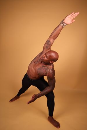 Muscular tattooed man in black leggings performing a side-angle yoga stretch in a warm-toned studio backdrop, barefoot with beaded bracelets — dynamic fitness portrait.
