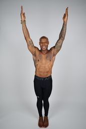 Smiling shirtless bald man with tattoos raises both arms overhead, wearing black compression leggings and a beaded bracelet, standing barefoot against a plain gray background.