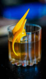 Close-up of a whiskey cocktail in a faceted rocks glass, topped with a zesty orange peel twist and resting on a dim bar surface