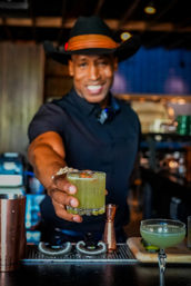 Smiling bartender in a black shirt and hat handing a bright green craft cocktail in a short faceted glass with a cherry garnish at a trendy cocktail bar