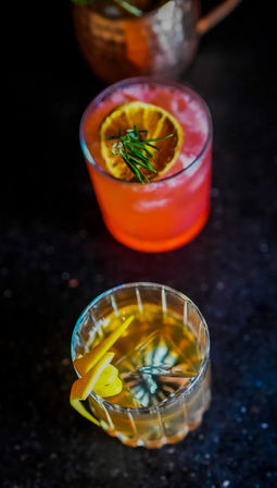 Overhead shot of two craft cocktails on a dark bar countertop — a pink citrus drink garnished with a dried orange slice and rosemary, and an amber old-fashioned with a lemon twist.