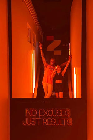Mirror selfie of two people posing on a stairwell in an orange-lit fitness studio with a neon motivational sign reading "NO EXCUSES JUST RESULTS" visible below