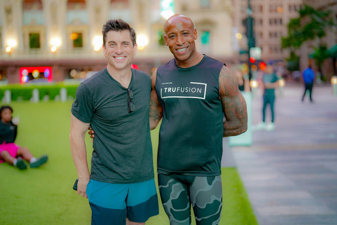 Two smiling men in workout clothes posing on a grassy downtown plaza at dusk, with city buildings and blurred lights in the background.