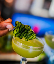 Close-up of a hand placing a curled cucumber ribbon garnish on a vibrant green cucumber cocktail in a coupe glass — fresh craft cocktail, mixology garnish