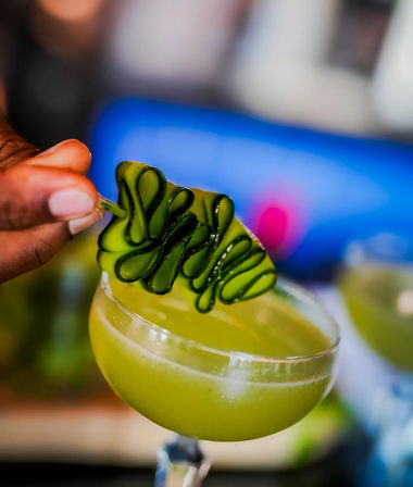 Close-up of a hand placing a curled cucumber ribbon garnish on a vibrant green cucumber cocktail in a coupe glass — fresh craft cocktail, mixology garnish