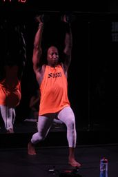 Barefoot athlete in an orange tank and white leggings doing an overhead dumbbell press from a lunge at a dim fitness studio with mirrored wall and water bottle nearby.