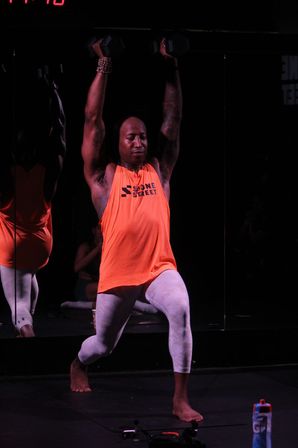 Barefoot athlete in an orange tank and white leggings doing an overhead dumbbell press from a lunge at a dim fitness studio with mirrored wall and water bottle nearby.