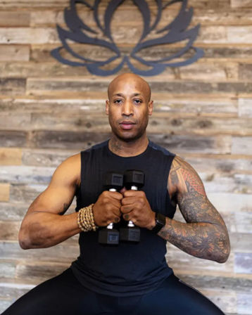 Fitness instructor holding a pair of dumbbells at chest level, tattooed arm visible, seated in front of a reclaimed wood wall with a lotus logo — studio workout portrait.