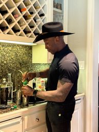 Stylish man in a black shirt and wide-brimmed hat mixing a cocktail at a home kitchen bar with a diamond wine rack and granite countertop