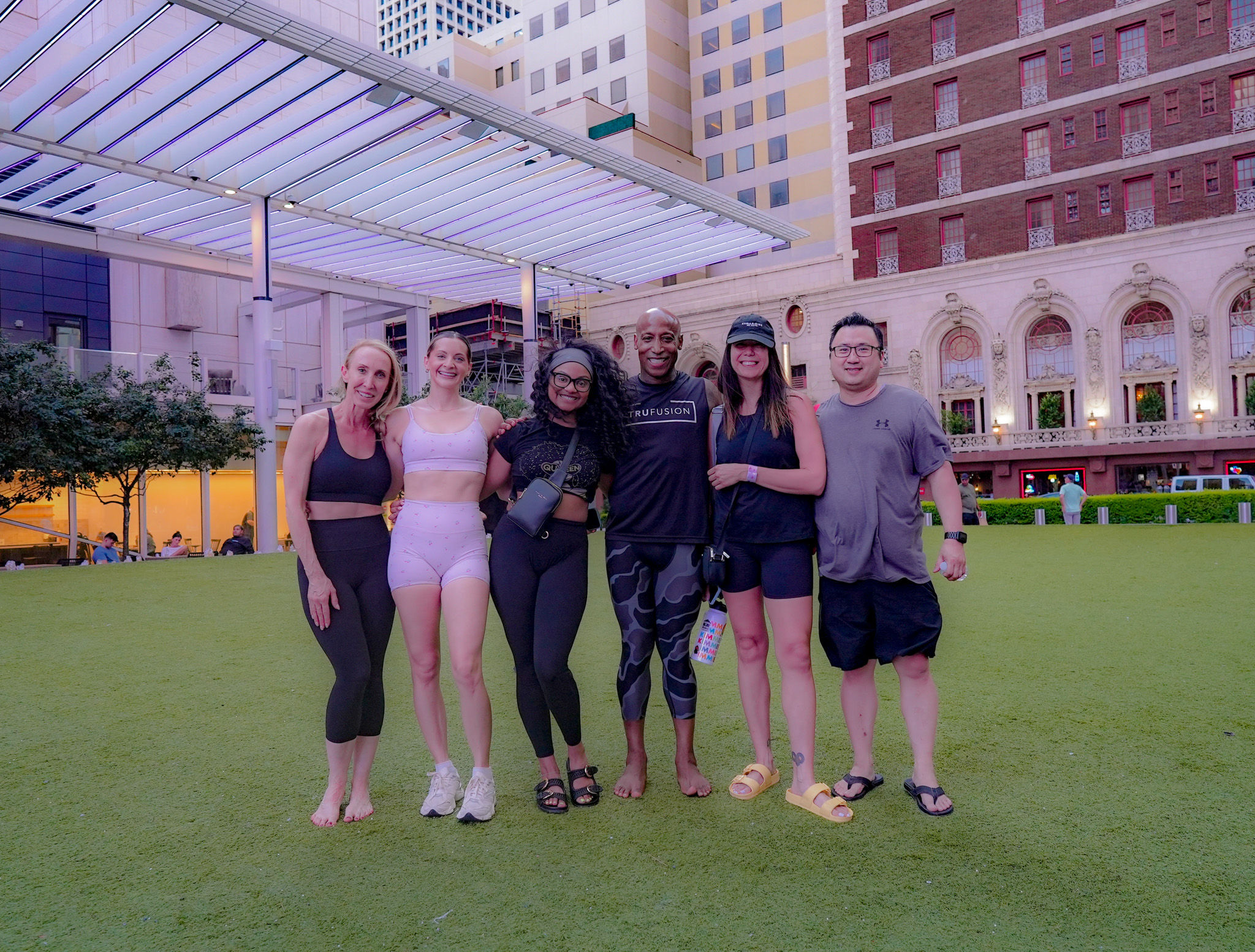 Six people in activewear smiling and posing on a downtown urban lawn beneath a modern lighted canopy and historic building facades — casual post-workout group photo in a city plaza.