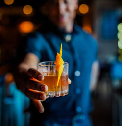 Bartender offering a whiskey cocktail in a rocks glass garnished with a bright orange twist in a cozy, dimly lit bar.