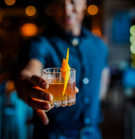 Bartender offering a whiskey cocktail in a rocks glass garnished with a bright orange twist in a cozy, dimly lit bar.