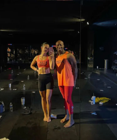 Two fit people in bright orange workout tops taking a mirror selfie inside a dim fitness studio, barefoot on black mats surrounded by water bottles and towels — post-workout class vibe.