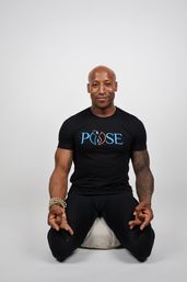 Smiling bald person kneeling on a meditation cushion in a neutral studio, wearing a black T-shirt with a colorful logo, hands in meditation mudra, beaded bracelet and tattooed arms.